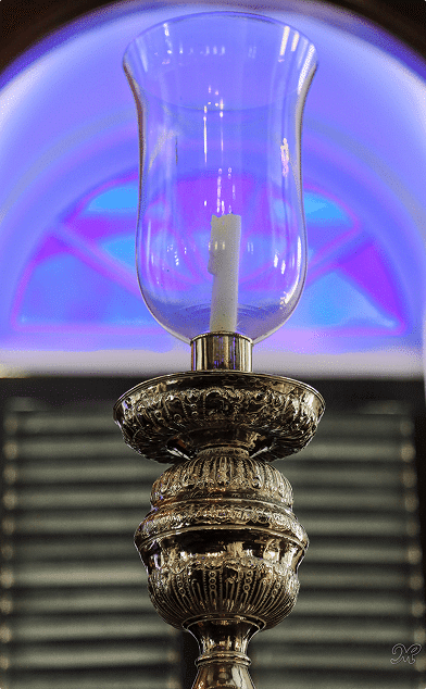 Ornate silver candlestick with a glass cover and blue arched stained glass window in the background.